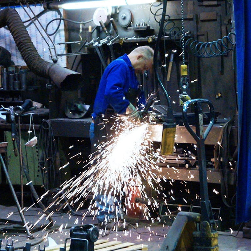 Arbeiter in Werkstatt schleift Metall an Werkbank, Funken sprühen in großem Bogen, umgeben von Werkzeugen, Hebezeug, Schläuchen und Werkstattausrüstung