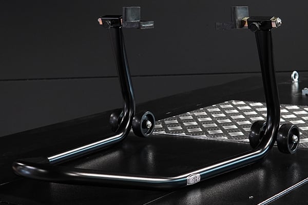 Black tubular motorcycle rear paddock stand with two upright arms, small wheels and padded lift points, placed on a diamond-plate platform against a dark background.