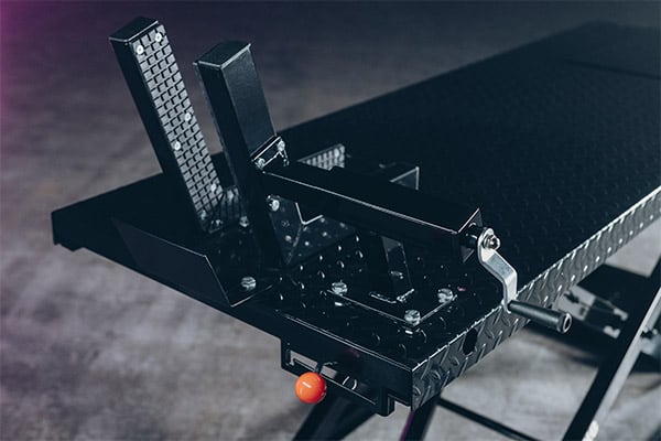 Soporte metálico negro para rueda de motocicleta montado en mesa elevadora con chapa diamantada, palanca de ajuste, tornillos visibles y botón naranja, enfoque en el mecanismo de sujeción