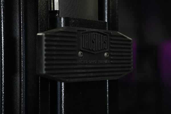Nahaufnahme eines schwarzen Vorhängeschlosses mit geriffelter Fläche und Markenprägung, an einem schwarzen Metallpfosten befestigt, dunkler unscharfer Hintergrund mit dezenten violetten Lichtpunkten
